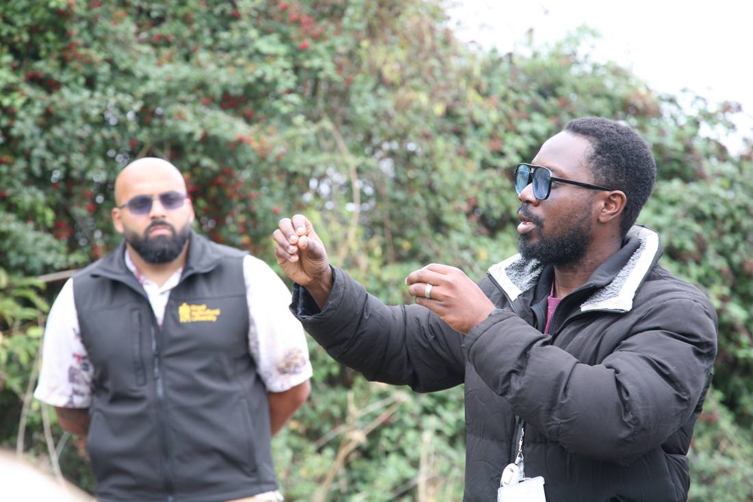 Emmanuel explaining something to a group outdoors beside a hedgerow